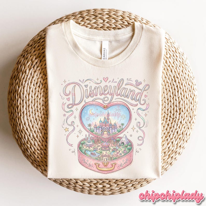 Może przedstawiać: Kremowa koszulka z pastelową ilustracją Disneylandu. Projekt zawiera pudełko w kształcie serca z zamkiem, fajerwerkami i napisem "Disneyland" w ozdobnej czcionce. Koszulka leży na tkanej powierzchni.