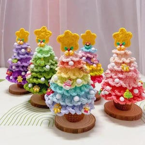 May include: Six colourful, crocheted Christmas trees are displayed. Each tree has a star topper, white bead accents, and small decorative ornaments. The trees are on small wooden bases, creating a festive holiday display.