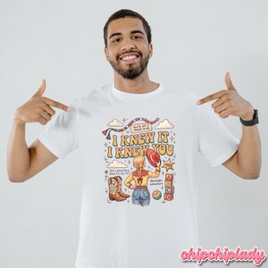 May include: A white t-shirt featuring a colourful graphic print with the text "I KNEW IT I KNEW YOU", "YOU'VE GOT A FRIEND IN ME", "It's a Jessie Story Baby Just Say Yet", and "nostalgic adventure". The design includes a cowgirl, boots, and blocks.