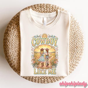 May include: A cream-colored t-shirt featuring a graphic print of two figures in cowboy hats walking down a path. The text "COWBOY LIKE ME" is printed in large, stylized letters above and below the illustration, which is framed by flowers and a sun.