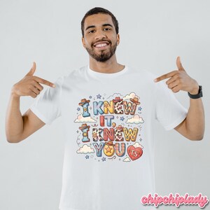 K&ouml;nnte beinhalten: Ein wei&szlig;es T-Shirt mit einem farbenfrohen Grafikdruck mit dem Text I KNEW IT, I KNEW YOU. Das Design enth&auml;lt verschiedene Muster, kleine Cowboyh&uuml;te, Wolken, Sterne und ein rotes Herz mit den Buchstaben TS im Inneren, alles in Blockform.