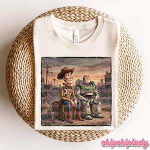 May include: Cream-colored t-shirt featuring a distressed graphic of Woody and Buzz Lightyear from Toy Story, sitting together. The image has a vintage, faded look, with a cityscape backdrop. The shirt has a classic crew neck and is made of a soft material.