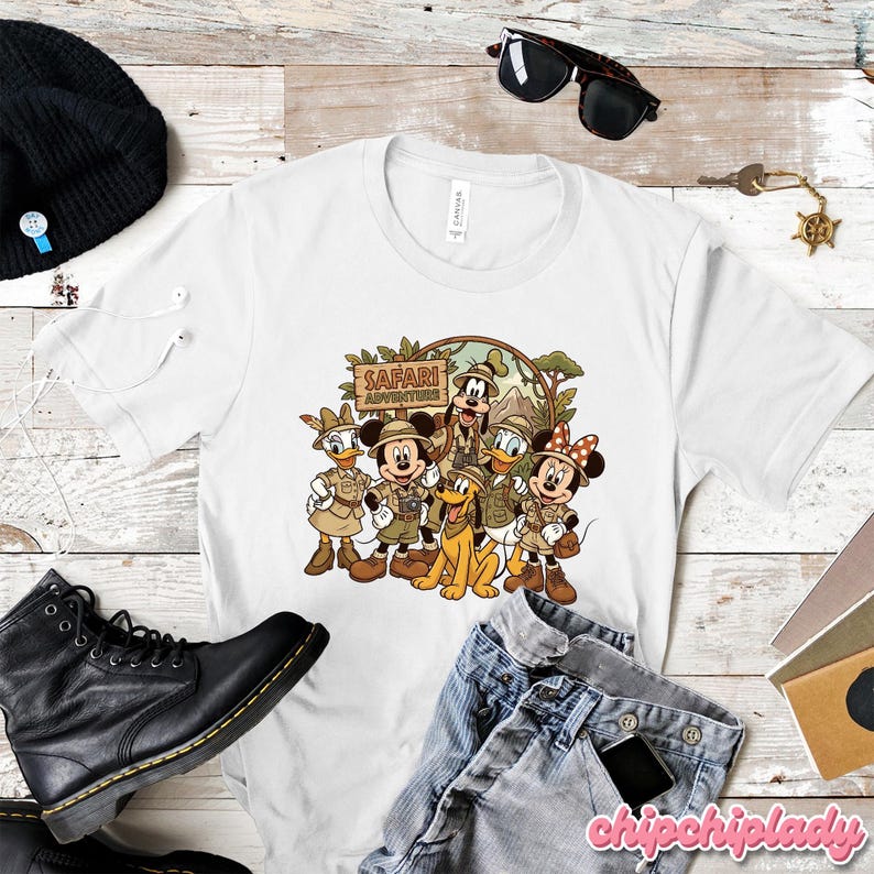 Puede incluir: Camiseta blanca con un dise&ntilde;o de personajes de Disney vestidos para un safari, incluyendo Mickey Mouse, Minnie Mouse, Donald, Daisy, Goofy y Pluto. El texto "Safari Adventures" aparece en un letrero de madera.