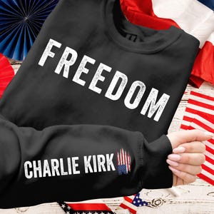 Peut inclure: Sweat-shirt noir avec le mot "FREEDOM" en grandes lettres blanches sur la poitrine. La manche porte l'inscription "CHARLIE KIRK" en blanc, avec un motif stylis&eacute; du drapeau am&eacute;ricain. Le sweat-shirt est pr&eacute;sent&eacute; sur une surface en bois avec des d&eacute;corations patriotiques.