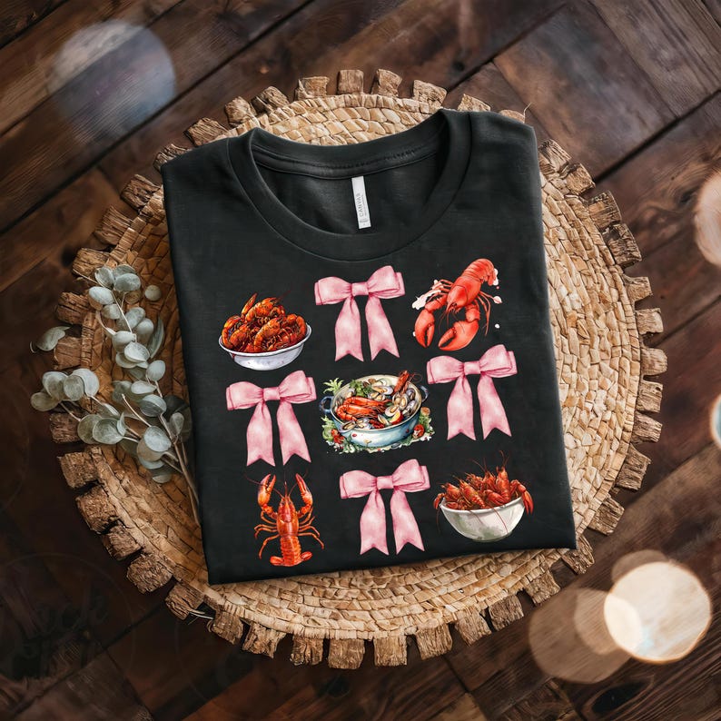 May include: Black t-shirt with a repeated pattern of pink bows and illustrations of crawfish, crawfish in bowls, and a pot of crawfish.