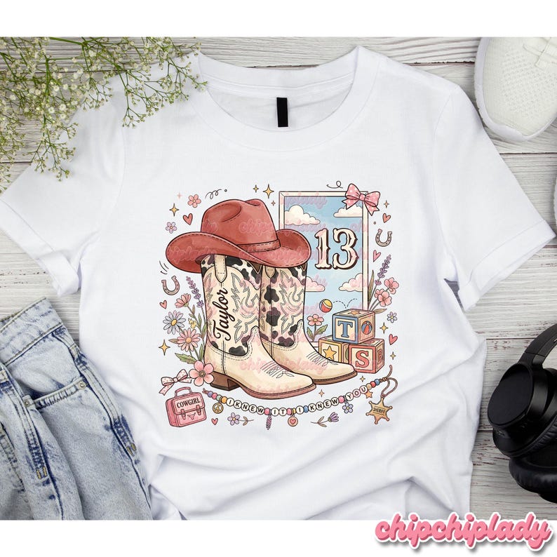Pode incluir: Uma t-shirt branca com uma impress&atilde;o gr&aacute;fica de um chap&eacute;u de cowboy rosa sobre botas western com o nome Taylor, o n&uacute;mero 13, blocos de alfabeto, uma pequena mala e um colar de contas com o texto I KNEW IT I KNEW YOU.