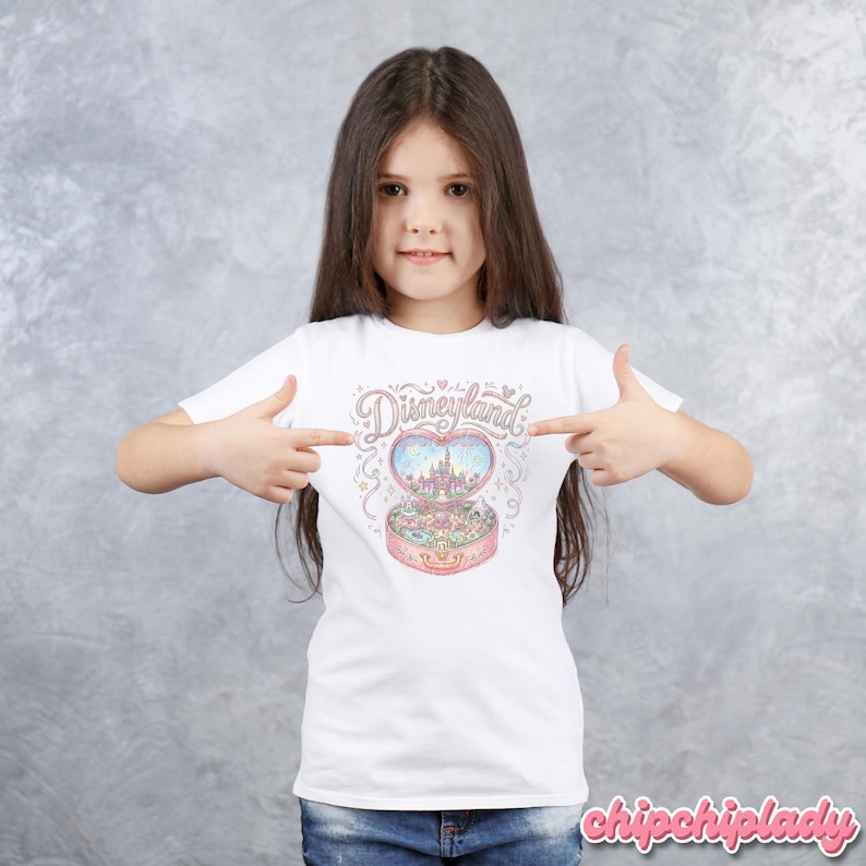 Może przedstawiać: Biała koszulka z kolorową ilustracją pozytywki Disneyland w kształcie serca. Projekt zawiera napis "Disneyland" w ozdobnej czcionce nad pozytywką. Pozytywka jest r&oacute;żowa i ma w środku zamek.