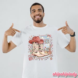Pode incluir: Uma t-shirt branca com uma impress&atilde;o gr&aacute;fica de um chap&eacute;u de cowboy cor-de-rosa sobre um par de botas western com padr&atilde;o de vaca. O design inclui o nome Taylor, o n&uacute;mero 13, blocos de letras, uma pequena mala, flores e estrelas.