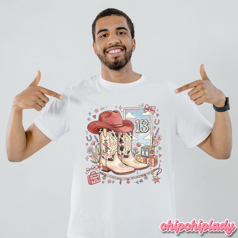 Pode incluir: Uma t-shirt branca com uma impress&atilde;o gr&aacute;fica de um chap&eacute;u de cowboy cor-de-rosa sobre um par de botas western com padr&atilde;o de vaca. O design inclui o nome Taylor, o n&uacute;mero 13, blocos de letras, uma pequena mala, flores e estrelas.