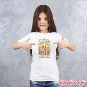May include: A white short-sleeved t-shirt featuring a colorful graphic print of two figures in cowboy hats walking through a field. The text "COWBOY LIKE ME" is printed in a decorative font above and below the illustration, with a sun icon at the top.