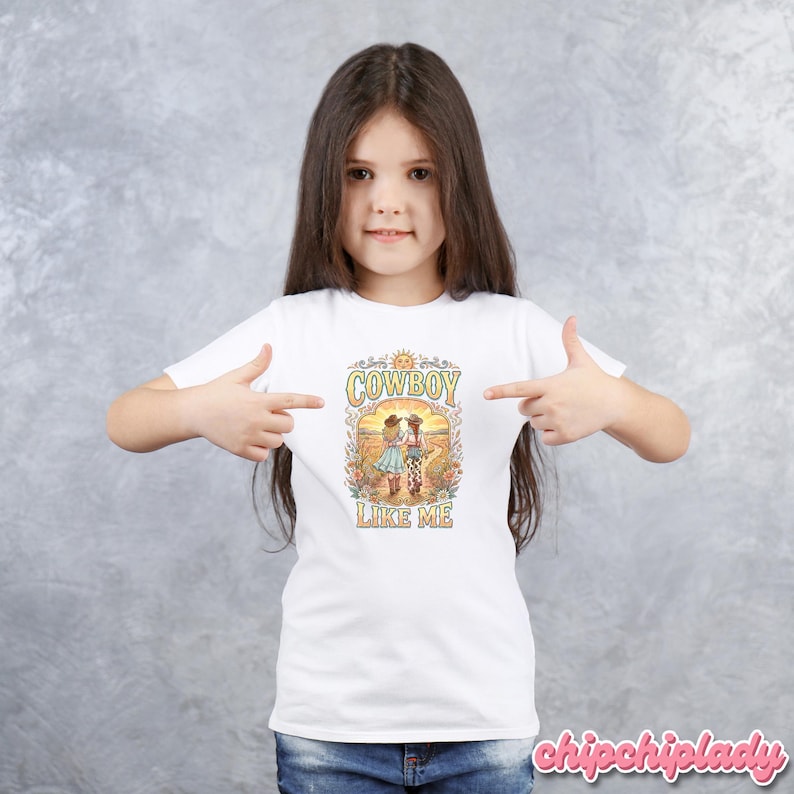 May include: A white short-sleeved t-shirt featuring a colorful graphic print of two figures in cowboy hats walking through a field. The text "COWBOY LIKE ME" is printed in a decorative font above and below the illustration, with a sun icon at the top.