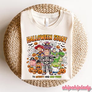 May include: Cream-colored t-shirt with a colorful Halloween-themed graphic. The design features characters from Toy Story, pumpkins, ghosts, and the text "HALLOWEEN STORY" and "TO INFINITY AND BOO-YOND!". The shirt is displayed on a woven surface.
