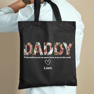 May include: Black tote bag with the word "DADDY" in white, filled with family photos. Below the word is the text "To the world you are one person but to us you are the world" with a heart and the name "Liam".