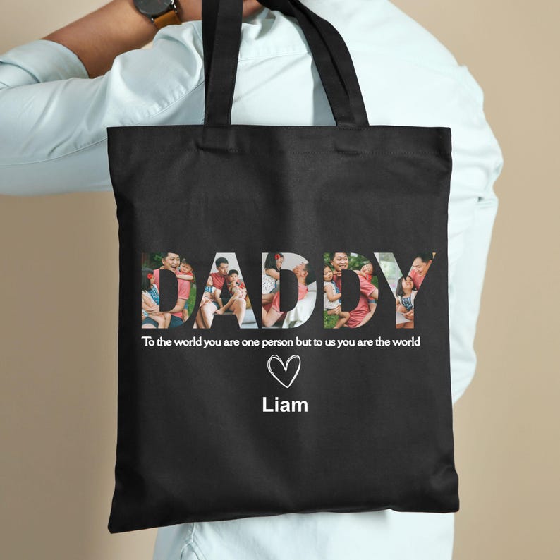 May include: Black tote bag with the word "DADDY" in white, filled with family photos. Below the word is the text "To the world you are one person but to us you are the world" with a heart and the name "Liam".