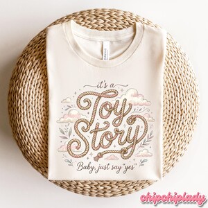 Puede incluir: Una camiseta de color crema con un dise&ntilde;o gr&aacute;fico que presenta el texto "it's a Toy Story" escrito en una fuente estilo cuerda, rodeado de ilustraciones de nubes rosas y hojas peque&ntilde;as. Debajo del texto principal, la frase "Baby, just say "yes"" est&aacute; impresa.