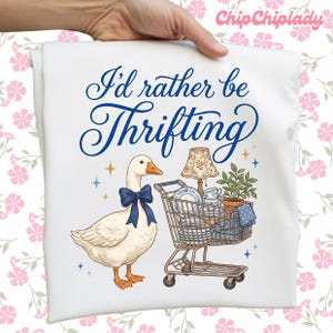 May include: White t-shirt with a whimsical illustration. A goose wearing a blue bow stands next to a shopping cart filled with vintage items, including a lamp and dishes. The text "I'd rather be Thrifting" is written in blue script.