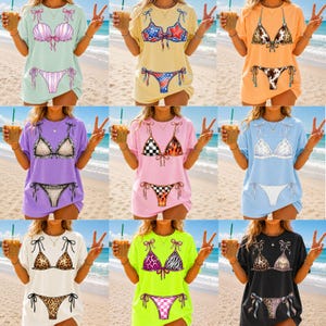 May include: A collection of nine t-shirts, each featuring a bikini top and bottom design. The shirts come in various colors, including light green, yellow, orange, purple, pink, blue, white, neon green, and black. The bikini designs vary, with patterns like seashells, stars, and animal prints.