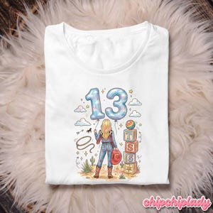 Pu&ograve; includere: Una t-shirt bianca con la grafica di una persona dai capelli biondi che indossa un completo in denim e stivali da cowboy. Il design include il numero 13 in carattere a palloncino blu, un lazo, cactus e una pila di blocchi con le lettere T, S, S.