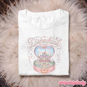 Może przedstawiać: Biała koszulka z sercowatym wzorem i napisem "Disneyland" w pastelowych kolorach. Serce otwiera się, ukazując miniaturową scenę Disneylandu z zamkiem, fajerwerkami i innymi atrakcjami. Tekst "chipchiplady" znajduje się na dole.