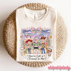 Peut inclure: T-shirt cr&egrave;me avec une illustration color&eacute;e de personnages de dessins anim&eacute;s populaires. Le motif comprend le texte "You've Got a Friend In Me" et des images de montagnes russes, de montgolfi&egrave;res et d'&eacute;toiles.