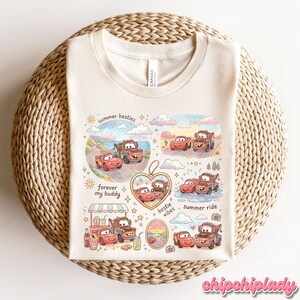 May include: Cream-colored t-shirt featuring a colorful graphic of cartoon cars in various scenes. The design includes text that reads "summer besties," "forever my buddy," and "bestie vibes." The shirt is laid on a woven surface.