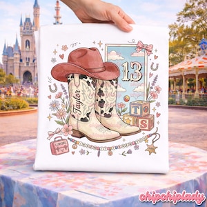 Pode incluir: Uma t-shirt branca com uma ilustra&ccedil;&atilde;o gr&aacute;fica de um chap&eacute;u de cowboy castanho sobre um par de botas de cowboy creme e pretas com padr&atilde;o de vaca. O design inclui o nome Taylor, o n&uacute;mero 13, blocos de letras, flores e o texto I KNEW IT I KNEW YOU.