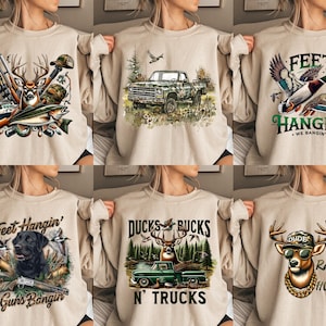 Op de afbeelding: Zes verschillende ontwerpen van beige sweatshirts met verschillende jacht- en natuurthema's. De ontwerpen omvatten een hert met geweien, eenden, een vrachtwagen, een hond en een bok. De tekst op de sweatshirts is "Feet Hangin' We Bangin'," "Ducks & Bucks N' Trucks," "Feet Hangin' Guns Bangin'," en "Dude Rather Be Hunting."