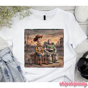 May include: White t-shirt featuring a graphic of Woody and Buzz Lightyear from Toy Story. Woody is wearing a cowboy hat and Buzz is in a spacesuit. The image has a distressed, vintage look.