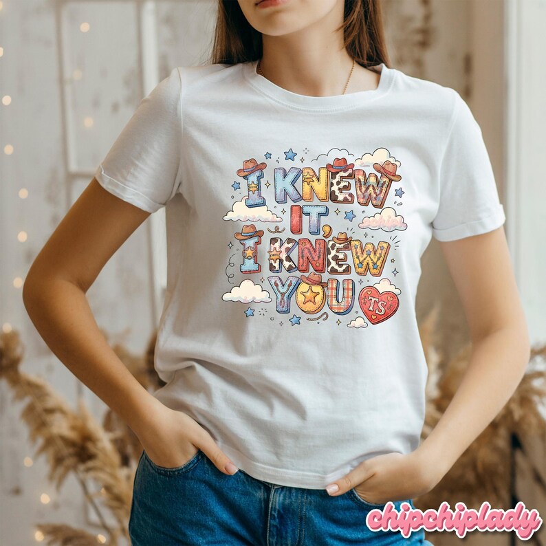 K&ouml;nnte beinhalten: Ein wei&szlig;es T-Shirt mit kurzen &Auml;rmeln und einem bunten Grafikdruck mit dem Text I KNEW IT, I KNEW YOU. Die Buchstaben sind mit Mustern wie Kuhflecken, Sternen und kleinen Cowboyh&uuml;ten verziert, begleitet von Wolken und einem roten Herz.