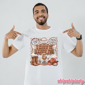 May include: A white t-shirt featuring a colorful graphic print with the text "I KNEW IT, I KNEW YOU", "5", "YOU'VE GOT A FRIEND IN ME", and "Infinity and Beyond". The design includes illustrations of cowboy boots, a cowboy hat, and alphabet blocks.