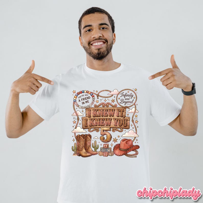 May include: A white t-shirt featuring a colorful graphic print with the text "I KNEW IT, I KNEW YOU", "5", "YOU'VE GOT A FRIEND IN ME", and "Infinity and Beyond". The design includes illustrations of cowboy boots, a cowboy hat, and alphabet blocks.