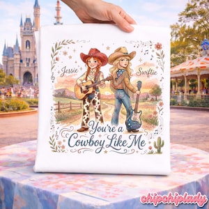 Pu&ograve; includere: Una t-shirt bianca con un'illustrazione grafica di due cowgirl con chitarre in un paesaggio rurale. Una &egrave; etichettata Jessie, l'altra Swiftie. Il testo recita You're a Cowboy Like Me, circondato da motivi floreali, stelle e note musicali.