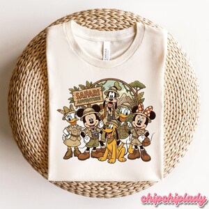Puede incluir: Camiseta color crema con un dibujo animado de personajes de Disney con atuendo de safari. Mickey Mouse, Minnie Mouse, Pato Donald, Daisy, Goofy y Pluto est&aacute;n representados con el texto "Safari Adventure".