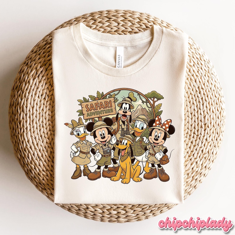 Puede incluir: Camiseta color crema con un dibujo animado de personajes de Disney con atuendo de safari. Mickey Mouse, Minnie Mouse, Pato Donald, Daisy, Goofy y Pluto est&aacute;n representados con el texto "Safari Adventure".
