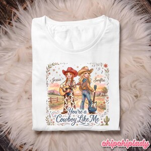 Pu&ograve; includere: Una t-shirt bianca con un'illustrazione grafica di due cowgirl con chitarre in un paesaggio rurale. Il testo recita Jessie, Swiftie e You're a Cowboy Like Me. Il design include accenti floreali, note musicali e un cactus.