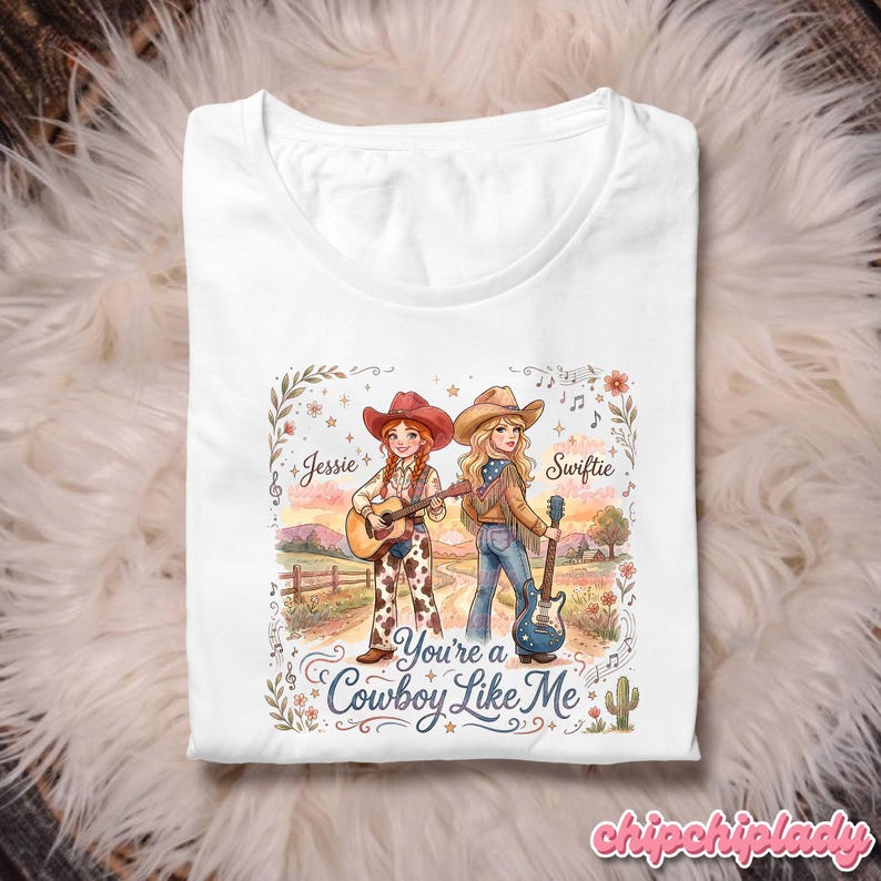 Pu&ograve; includere: Una t-shirt bianca con un'illustrazione grafica di due cowgirl con chitarre in un paesaggio rurale. Il testo recita Jessie, Swiftie e You're a Cowboy Like Me. Il design include accenti floreali, note musicali e un cactus.