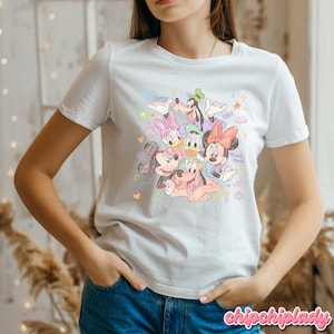 May include: White t-shirt featuring a colorful illustration of Disney characters including Mickey Mouse, Minnie Mouse, Donald Duck, Daisy Duck, Goofy, and Pluto. The design includes the words "Minnie Mouse" and "Goofy".