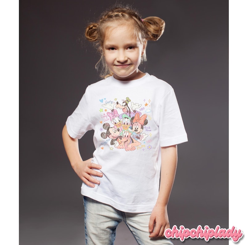 May include: A white t-shirt featuring a colorful graphic of Disney characters, including Mickey Mouse, Minnie Mouse, Donald Duck, Daisy Duck, Goofy, and Pluto. The shirt is short-sleeved and worn by a young person.