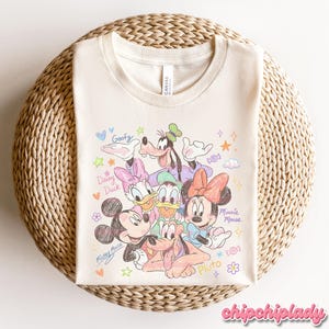 May include: Cream-colored t-shirt featuring a colorful sketch of Disney characters Mickey Mouse, Minnie Mouse, Donald Duck, Daisy Duck, Goofy, and Pluto. The shirt has the word "Canvas" printed on the tag.