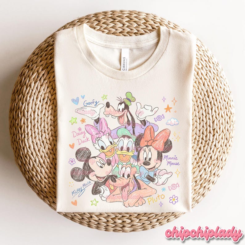 May include: Cream-colored t-shirt featuring a colorful sketch of Disney characters Mickey Mouse, Minnie Mouse, Donald Duck, Daisy Duck, Goofy, and Pluto. The shirt has the word "Canvas" printed on the tag.