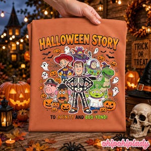 May include: Orange Halloween-themed t-shirt featuring a cartoon graphic with the text "HALLOWEEN STORY" and "TO INFINITY AND BOO-YOND!". The graphic includes various characters, pumpkins, ghosts, and bats. The shirt is held up by a hand.