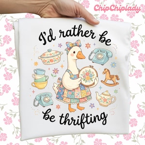 May include: White t-shirt with a whimsical illustration of a goose wearing a floral bow and apron, holding a floral embroidery hoop. The text reads "I'd rather be thrifting" in black script, surrounded by vintage-style illustrations.