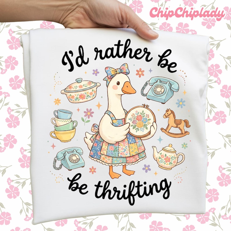 May include: White t-shirt with a whimsical illustration of a goose wearing a floral bow and apron, holding a floral embroidery hoop. The text reads "I'd rather be thrifting" in black script, surrounded by vintage-style illustrations.