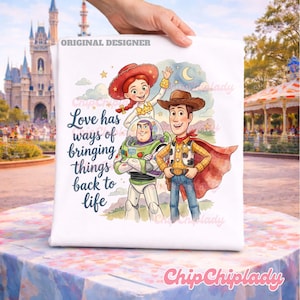 May include: A white t-shirt featuring a colorful illustration of Jessie, Buzz Lightyear, and Woody from Toy Story. The text reads, Love has ways of bringing things back to life. The design includes a crescent moon, stars, and clouds in a watercolor style.