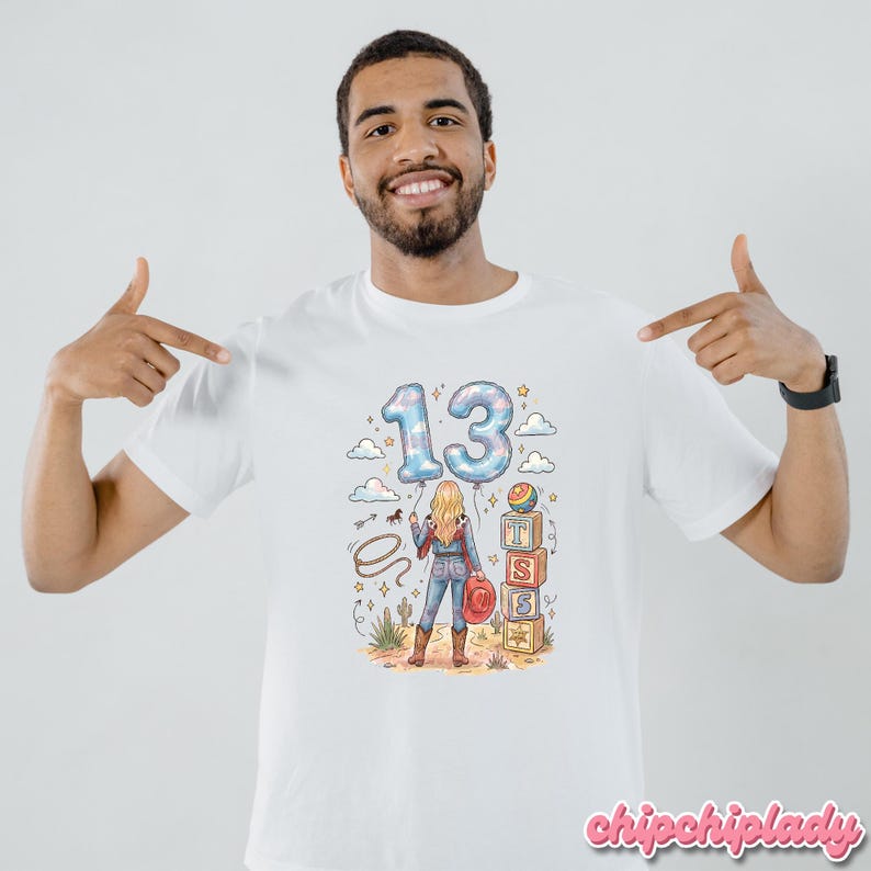 Pu&ograve; includere: Una t-shirt bianca con un'illustrazione grafica di una persona in un paesaggio desertico sotto il numero 13 a forma di palloncino. Il design include un lazo, cactus, una pila di blocchi con le lettere T, S, S e un cappello da cowboy.