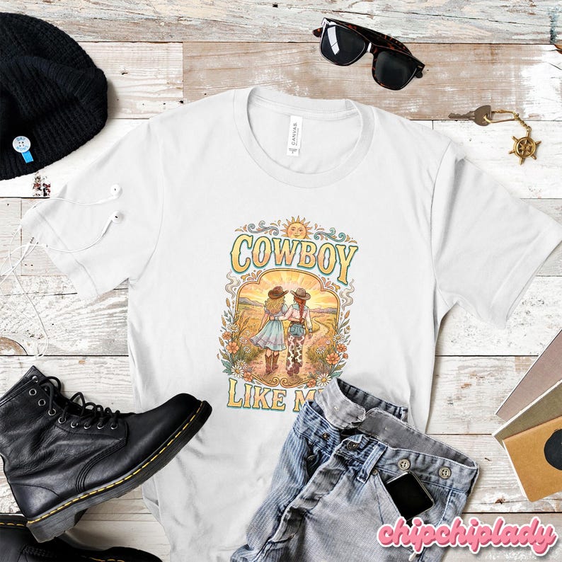 May include: A white short-sleeve t-shirt featuring a graphic print of two people in cowboy hats walking through a field. The text "COWBOY LIKE ME" is printed in yellow and blue above and below the illustration, which includes a sun and flowers.