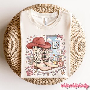 Pode incluir: Uma t-shirt cor de creme com uma impress&atilde;o gr&aacute;fica de um chap&eacute;u de cowboy rosa, uma bota de cowboy com padr&atilde;o de vaca com o nome Taylor, o n&uacute;mero 13, blocos de alfabeto, uma pequena mala rosa com a etiqueta Cowgirl e um colar de contas com o texto I KNEW IT I KNEW YOU.