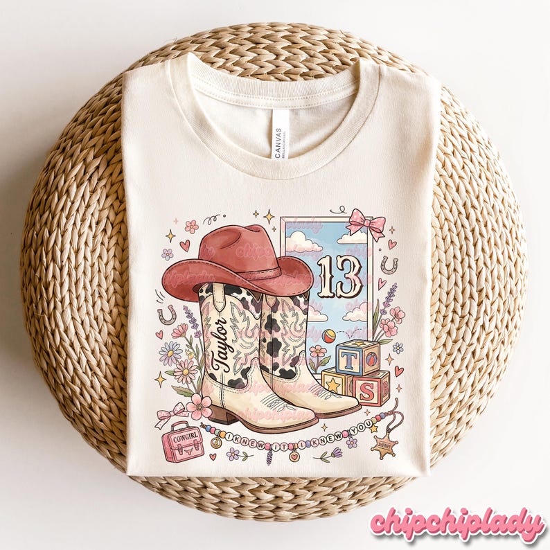 Pode incluir: Uma t-shirt cor de creme com uma impress&atilde;o gr&aacute;fica de um chap&eacute;u de cowboy rosa, uma bota de cowboy com padr&atilde;o de vaca com o nome Taylor, o n&uacute;mero 13, blocos de alfabeto, uma pequena mala rosa com a etiqueta Cowgirl e um colar de contas com o texto I KNEW IT I KNEW YOU.