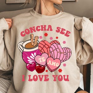 Op de afbeelding: Een beige sweatshirt met een grafisch ontwerp met een kopje warme chocolademelk, kaneelstokjes en hartvormige gebakjes. De tekst "Concha See" staat bovenaan en "Te Amo I Love You" onderaan.