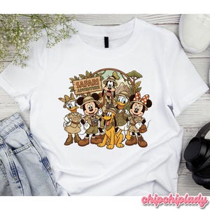 Puede incluir: Camiseta blanca con un gr&aacute;fico colorido de personajes de Disney con atuendo de safari. El dise&ntilde;o incluye Mickey Mouse, Minnie Mouse, Donald Duck, Daisy Duck, Goofy y Pluto, con las palabras "Safari Adventure" en un letrero de madera.